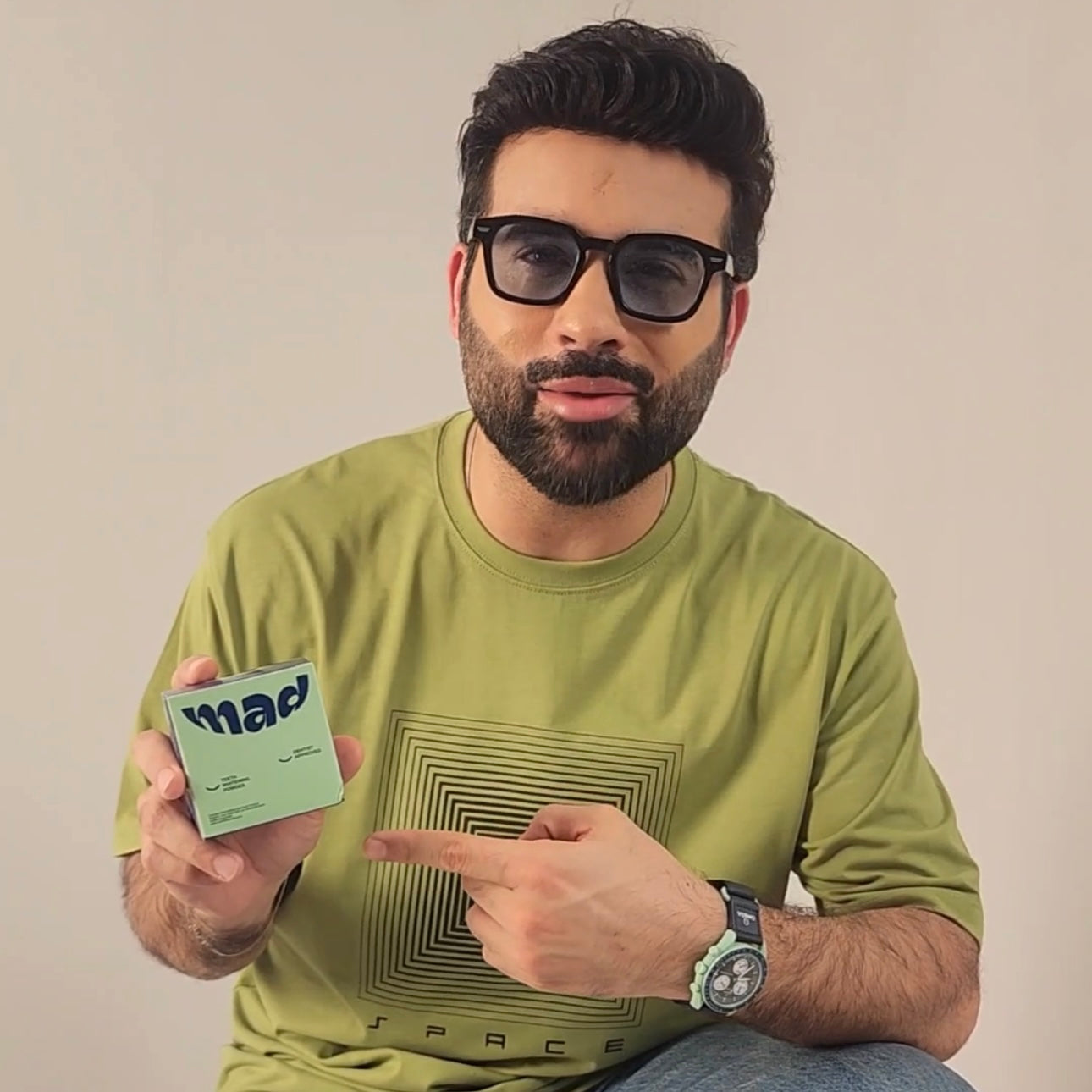 Man wearing glasses and a green shirt holding a product with a brand logo. Mad Care Teeth Whitening Powder 